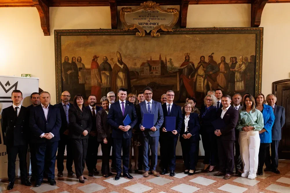 Grupa osób, głównie w garniturach, pozuje do pamiątkowego zdjęcia w pomieszczeniu historycznym z dużym obrazem na ścianie; trzech mężczyzn w pierwszym rzędzie t