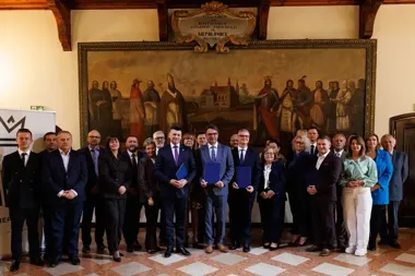 Grupa osób, głównie w garniturach, pozuje do pamiątkowego zdjęcia w pomieszczeniu historycznym z dużym obrazem na ścianie; trzech mężczyzn w pierwszym rzędzie t