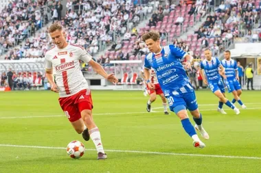 Piłkarz w biało-czerwonym stroju prowadzi piłkę przy linii boiska, a zawodnik w niebieskim stroju go dogania podczas meczu ligowego na stadionie z widzami.